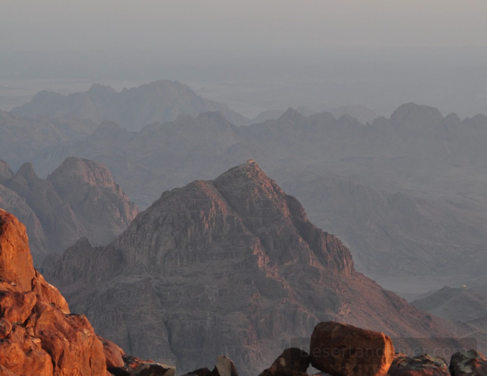 Sunrise view from Katherine Mountain in the Sinai Peninsula, looking over layered desert mountains and rocky foreground in warm light.