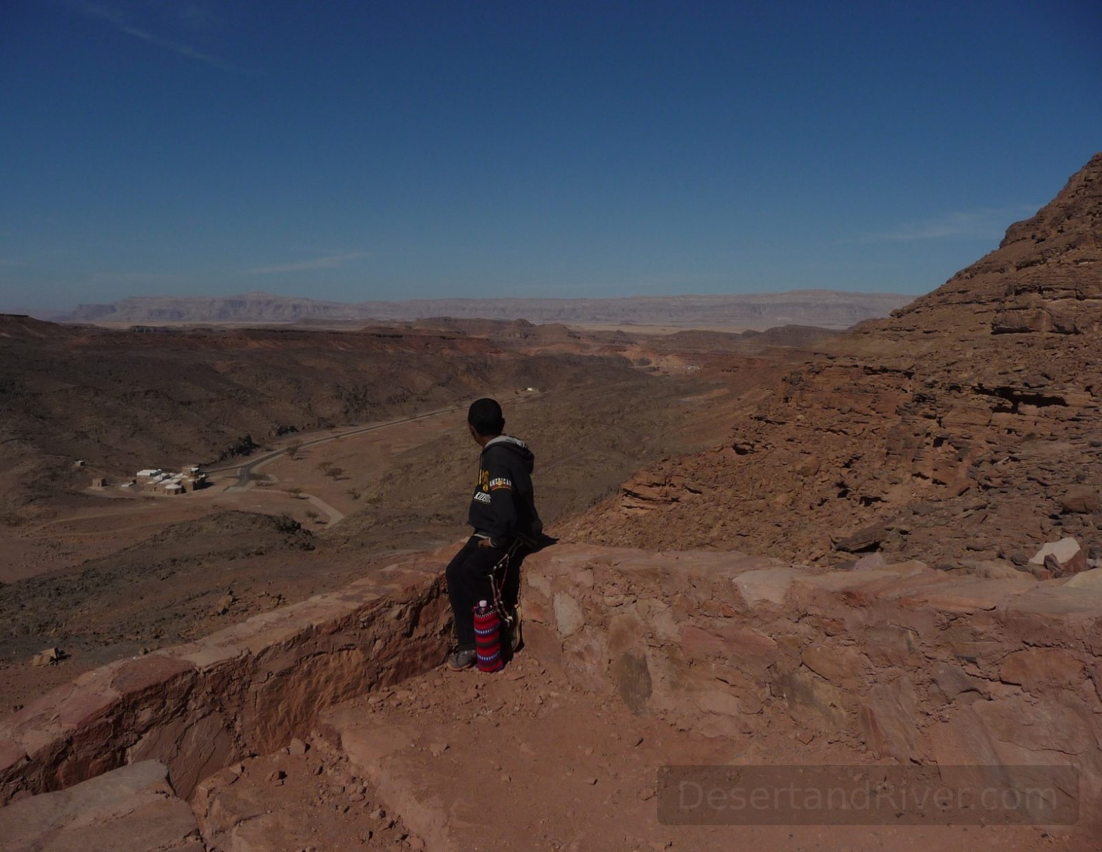View over Rod el-Eir wadi in the Sinai Peninsula, Egypt, with a Bedouin guide sitting on a rocky ledge above the desert valley.